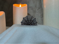 A black spiderweb-shaped brooch embellished with sparkling clear stones, displayed on a white fabric surface with glowing candles in the background.