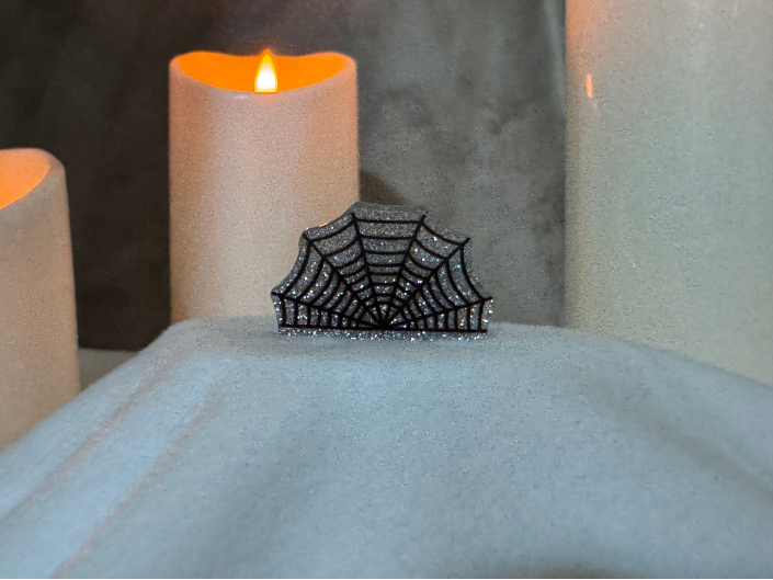 A black spiderweb-shaped brooch embellished with sparkling clear stones, displayed on a white fabric surface with glowing candles in the background.