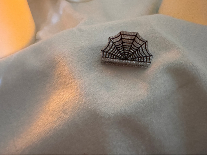 A small spiderweb-shaped brooch or pin with a dark metallic web pattern, displayed on a soft, light gray fabric surface with warm, yellow-toned lighting.
