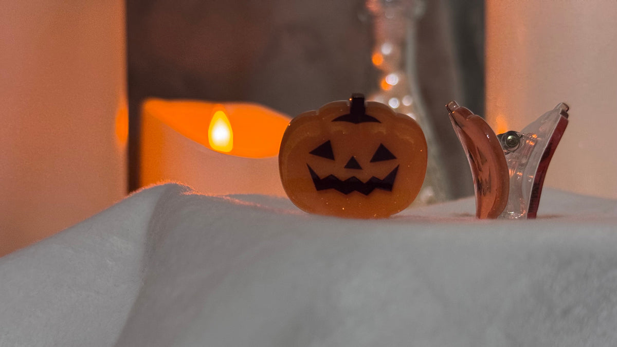 A small orange jack-o'-lantern hair clip with a carved black face sits on a white fabric-covered surface, with a warm glowing candle blurred in the background.