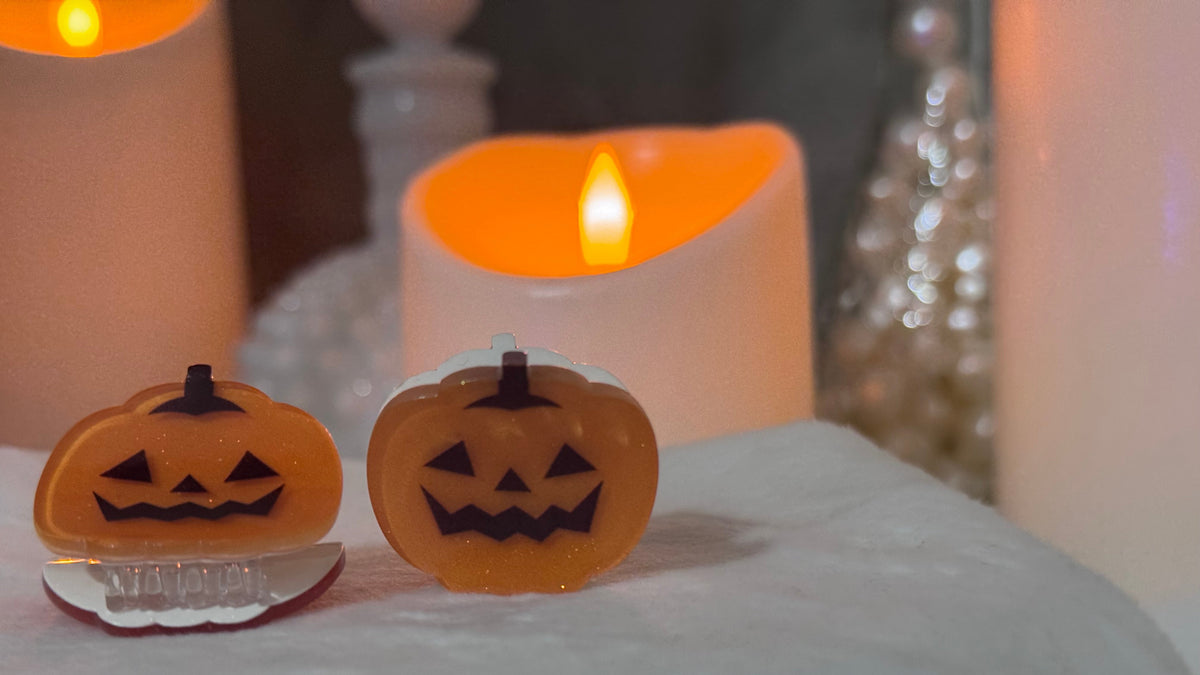 Two small jack-o'-lantern-shaped hair clips with carved pumpkin faces, displayed on a white fabric surface with glowing candles in the background.