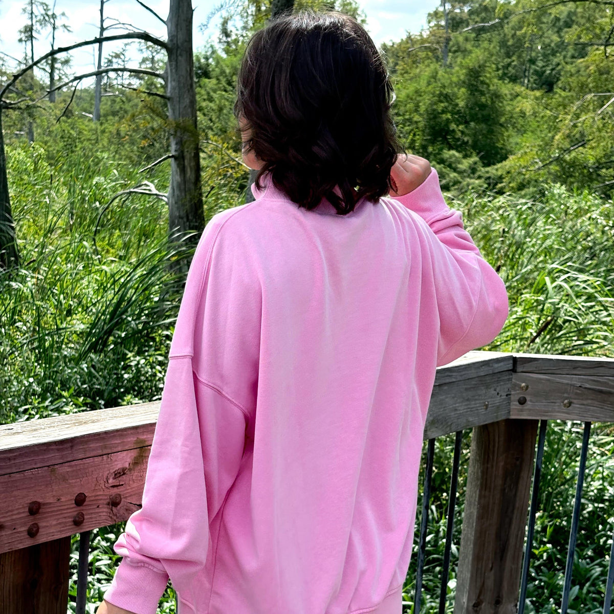 A person viewed from behind wearing an oversized light pink sweatshirt and denim shorts, standing on a wooden deck with a lush green outdoor background.
